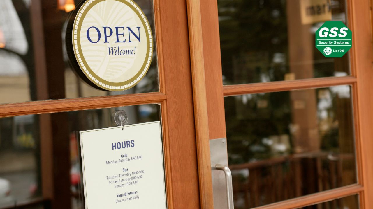 A wooden-framed glass door with an "OPEN" sign and business hours displayed, reflecting the street outside. The "GSS Security Systems" logo is in the top right corner.