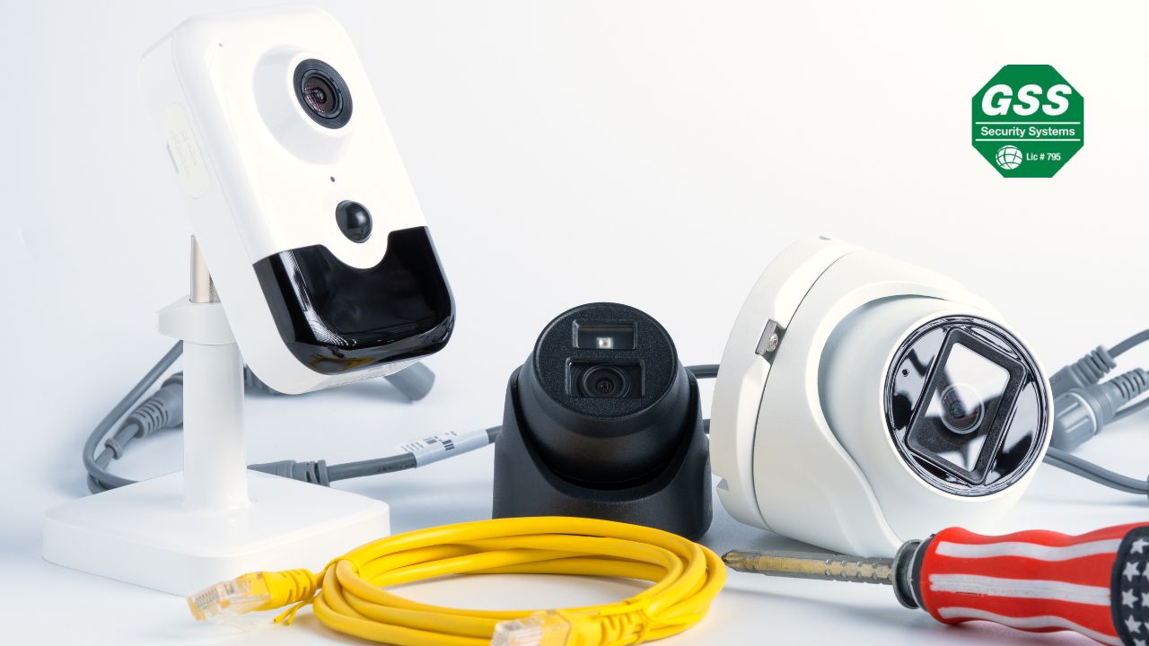 Various types of security cameras and equipment, including a white indoor camera, a black dome camera, a white dome camera, a yellow Ethernet cable, and a screwdriver, displayed on a light background. The logo of GSS Security Systems is visible in the top right corner, representing a focus on home security and alarm system setups.