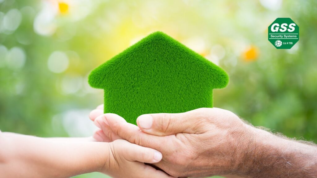 Hands of an adult and child holding a green, grass-textured house cutout against a blurred natural background with sunlight. The GSS Security Systems logo is displayed in the top right corner, symbolizing a focus on sustainable living and environmentally friendly home practices.