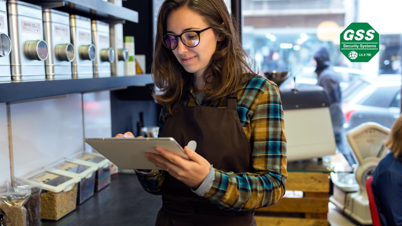 A young woman wearing glasses and an apron uses a tablet inside a cozy coffee shop. The "GSS Security Systems" logo is displayed in the top right corner.