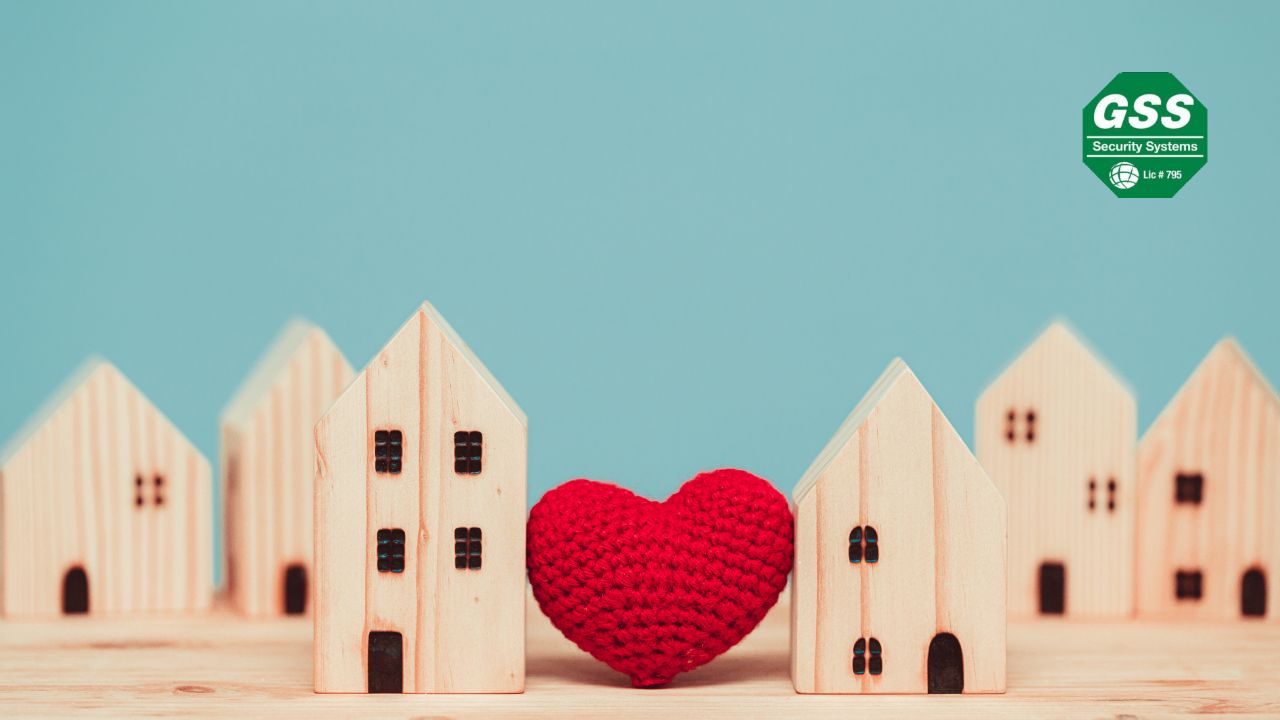 A group of small wooden houses is arranged on a wooden surface, with a bright red knitted heart placed between two of them. The background is a soft blue, and the "GSS Security Systems" logo is displayed in the top right corner.